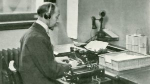 Man wearing suit and tie sits at a desk in front of a sunny window, typing on a manual typewriter. A cord from a machine on a table beside him runs to a pair of headphones on his head.