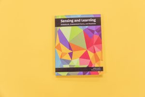 Sensing and Learning Guidebook, Assessment Forms, and Routines resting on a yellow background. The cover of the book showcases variations of pomegranate, teal, gold, and green.