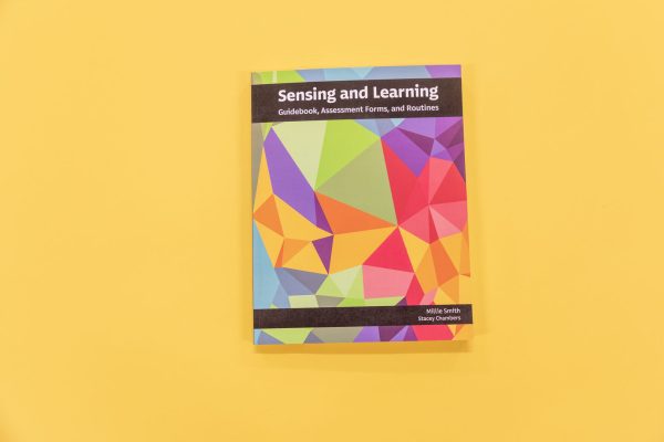Sensing and Learning Guidebook, Assessment Forms, and Routines resting on a yellow background. The cover of the book showcases variations of pomegranate, teal, gold, and green.