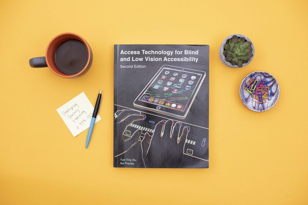 Image of the book, Utilizing Access Technology for Blind and Low Vision Accessibility in the Evaluation Process, on a table surrounded by a coffee cup and a plant.