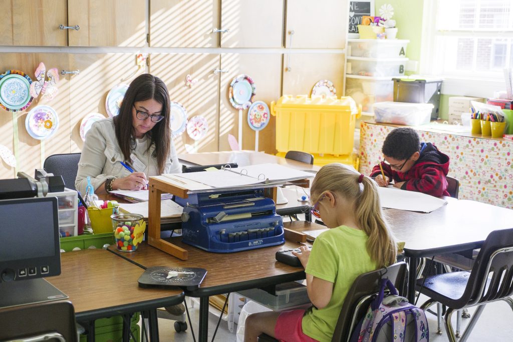 A teacher and two young students sit around a table that has a blue Perkins brailler on top of it. One student is writing on a piece of paper, one student is using a refreshable braille display, and the teacher is writing in a notebook.