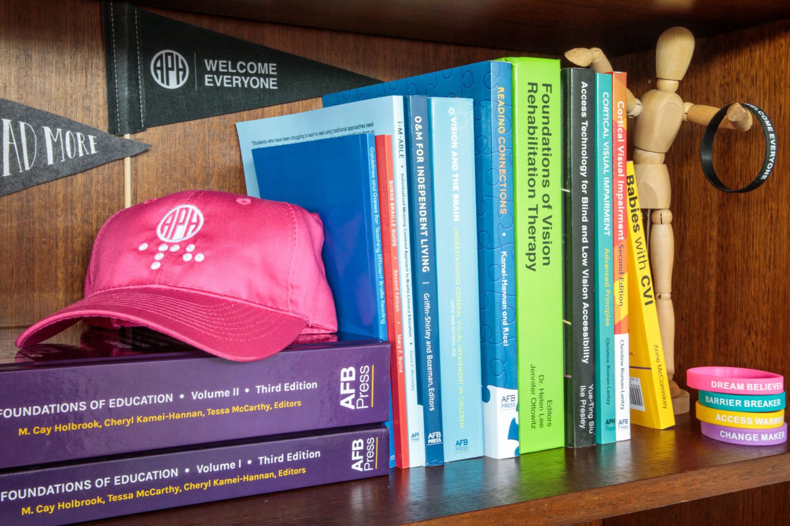 A collection of APH Press books, including Reading Connections: Strategies for Teaching Students with Visual Impairments, sit on a bookshelf alongside a pink hat with the APH logo on it, a small flag that has the APH logo and the words "welcome everyone" on it, a stack of APH branded bracelets, and a wooden drawing model of a person.
