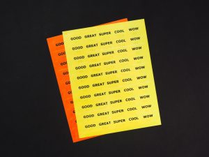 Two sheets of Feel-n-Peel stickers stacked on top of each other. The yellow sticker sheet resting on top of the orange sticker sheet, shows the following stickers with print and braille labels: Good, Great, Super, Cool, and Wow.