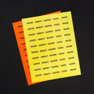 Two sheets of Feel-n-Peel stickers stacked on top of each other. The yellow sticker sheet resting on top of the orange sticker sheet, shows the following stickers with print and braille labels: Good, Great, Super, Cool, and Wow.