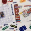 A close-up view of the Snap Circuits Jr. 130 Access Kit. The clear breadboard is displaying an exercise that uses the green power switch, one blue # 3 connector piece, a red fan, and two AA batteries inside a clear battery holder. The user guide is featured to the right of the breadboard