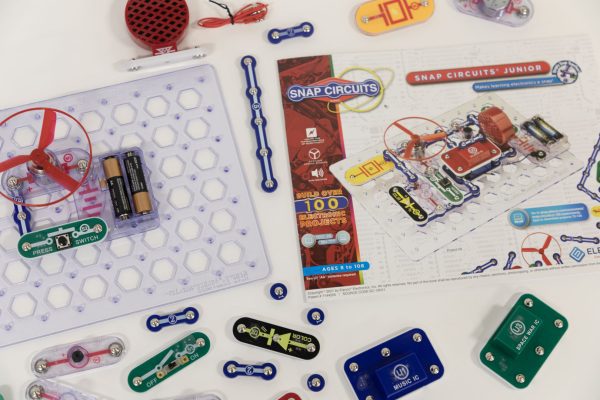 A close-up view of the Snap Circuits Jr. 130 Access Kit. The clear breadboard is displaying an exercise that uses the green power switch, one blue # 3 connector piece, a red fan, and two AA batteries inside a clear battery holder. The user guide is featured to the right of the breadboard
