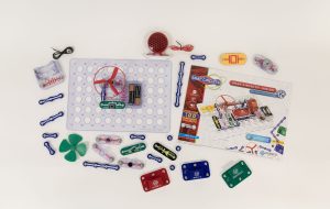 Each Snap Circuits Jr. 130 Access Kit part is displayed over a white background. An exercise is built on the breadboard showcasing the green power switch, one blue # 3 connector piece, a red fan, and two AA batteries inside a clear battery holder.