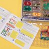The Snap Circuits Jr. 130 Access Kit user guide is flipped open to display Project 1 Electric Light, Project 2 Color Light, Project 4 Flying Saucer, and Project 6 Space War on top of a yellow background. The clear breadboard is displaying an exercise that uses the green power switch, one blue # 3 connector piece, a red fan, and two AA batteries inside a clear battery holder. A portion of the kit organized in the box is displayed in the upper left-hand corner.