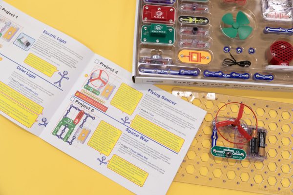 The Snap Circuits Jr. 130 Access Kit user guide is flipped open to display Project 1 Electric Light, Project 2 Color Light, Project 4 Flying Saucer, and Project 6 Space War on top of a yellow background. The clear breadboard is displaying an exercise that uses the green power switch, one blue # 3 connector piece, a red fan, and two AA batteries inside a clear battery holder. A portion of the kit organized in the box is displayed in the upper left-hand corner.