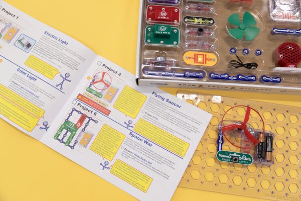 The Snap Circuits Jr. 130 Access Kit user guide is flipped open to display Project 1 Electric Light, Project 2 Color Light, Project 4 Flying Saucer, and Project 6 Space War on top of a yellow background. The clear breadboard is displaying an exercise that uses the green power switch, one blue # 3 connector piece, a red fan, and two AA batteries inside a clear battery holder. A portion of the kit organized in the box is displayed in the upper left-hand corner.