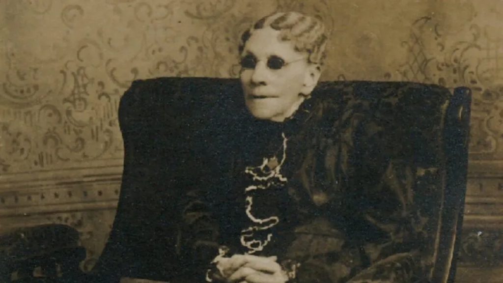 Portrait of a very old woman sitting in an armchair. Her thinning hair is fixed in waves pulled back from her forehead. She wears a black dress with white lace cascading from the collar to the waist.