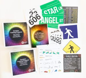 Envision Kit without Optical Aids opened to show training curriculum, activity cards, signage, restaurant menu, and additional large print resources.