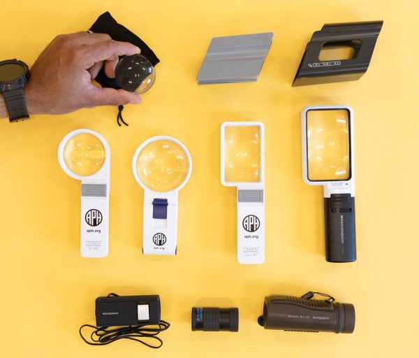 Optical aids including magnifiers and magnifier holders are shown out of box over a yellow background. In the top left-hand corner, a hand is holding a circular magnifier.