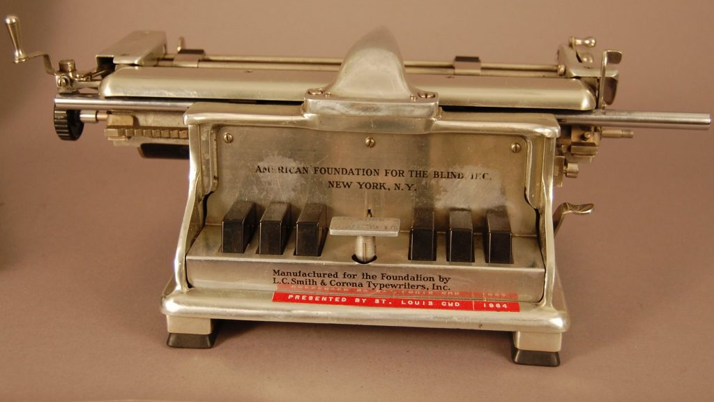 Braillewriter made of cast aluminum alloy, highly polished on the outside. It has piano-like keys in the front, separated by a stainless-steel space bar. Two folding arms extend from the back to hold the paper.