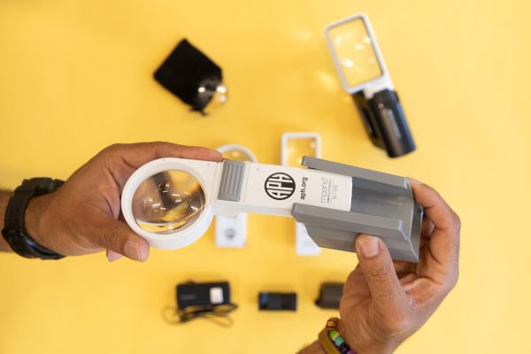 Multiple magnifiers and magnifier holders resting inside the black case included in the kit. 4. Placing-Magnifier-in-holder – Two hands focused in on holding a white circular magnifier that is being slid into a gray magnifier holder. There are additional optical aids resting over a yellow background.