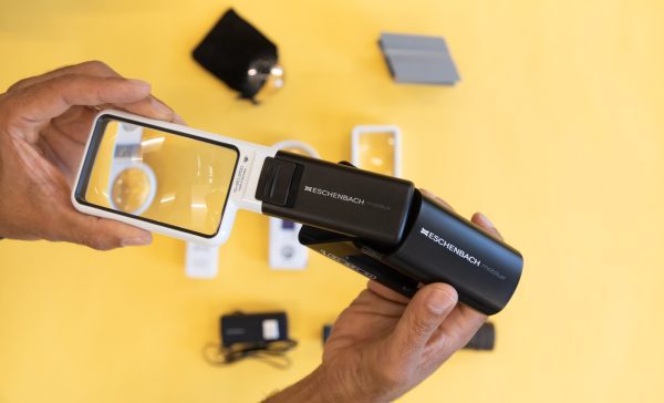 Two hands focused in on holding a white rectangular magnifier that is being slid into a black magnifier holder. There are additional optical aids resting over a yellow background.