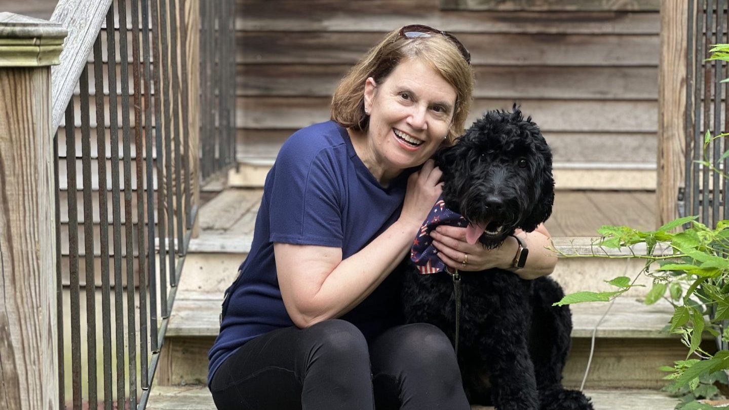 A woman smiles and closely holds a dog with curly black fur as they sit on wooden porch steps together.