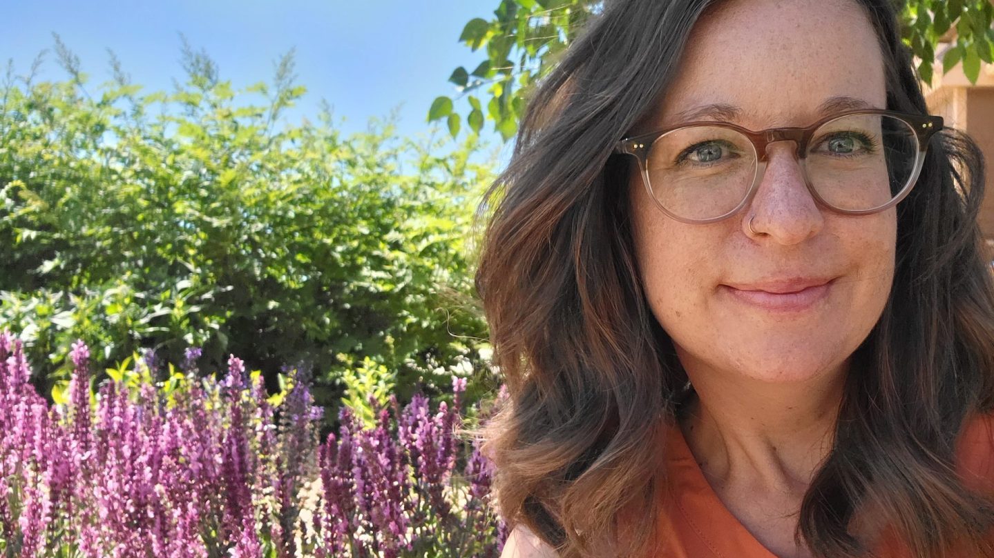 A woman with brown hair, glasses, and an orange shirt smiles in front of some tall, thin purple flowers, some greenery, and a blue sky.