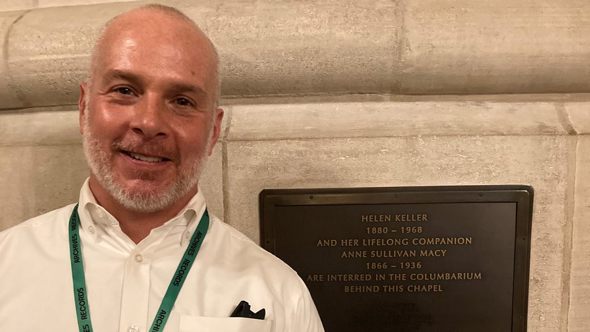Head-and-shoulders photo of a man wearing a white shirt with a lanyard around his neck. Beside him, fixed to the wall, is a bronze plaque. It reads: Helen Keller, 1880-1968, and her lifelong companion Anne Sullivan Macy, 1866-1936, are interred in the columbarium behind this chapel.