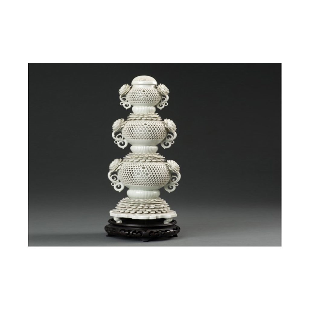 White porcelain incense burner, about 15 inches tall. Three bowls, perforated with a honeycomb design, sit on top of each other. Each bowl has two handles; each handle has a multi-petal flower design at the top.