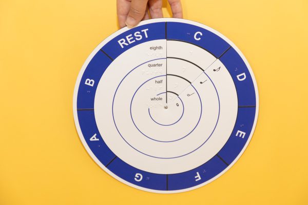 The Music Braille Wheel has a blue outer ring and four white inner rings displayed over a yellow background. The wheel consists of five rings or sections in total. The outer most section shows the notes: C, D, E, F, G, A, B, and rest in large print and braille. The inner rings in order from outward to inward show print and braille for eighth, quarter, half, and whole notes in print and braille for each of the notes listed in the outer ring.
