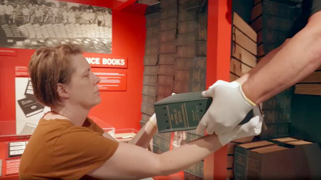 A woman dressed in a brown T-shirt accepts a braille book being handed to her. Behind her, on exhibit, are dozens of other books. The words “The World Book Encyclopedia” are written on the book’s spine.