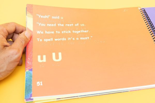 A hand holding an orange open page of Alphabet Scramble that shows large print text in line with braille.