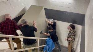 Five men wrestle with a large rectangular tan metal cabinet in a stairway. The stairsteps and handrails are covered with cardboard and packing blankets. Two men above the cabinet tilt it backwards, while two others below extend their arms to arrest its downward motion. A fifth man on the landing below gestures, as if shouting instructions or a warning.
