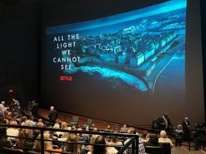 A large group of people sit in a curved seating area in front of a gigantic movie screen displaying a World War II era town on the coast and the text: "All the Light We Cannot See" and the Netflix logo.