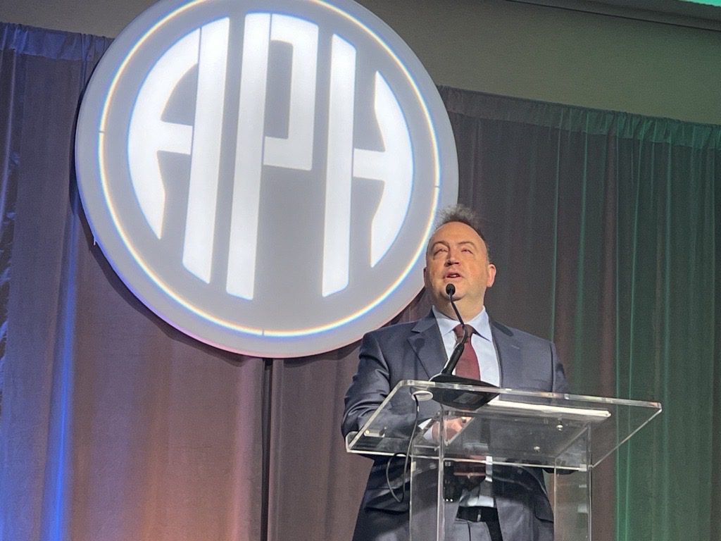 NFB President Mark Riccobono speaks into a microphone from behind a clear glass podium. The APH logo is projected behind him.