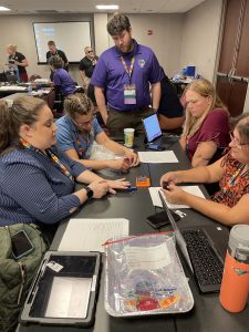 Four women sitting at a table and a man standing at the end of the table converse as they work with Snap Circuits parts.