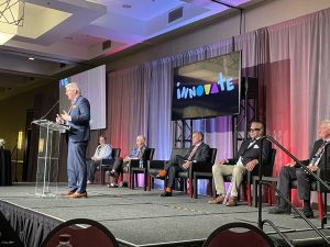 APH President Craig Meador speaks from a low stage. Behind him are two women and three men in business wear sitting on black chairs and a screen with the 2023 Annual Meeting logo on it, which is the word “innovate.”