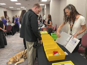 A woman holds open a white binder, showing it to a man with a guide dog. On the table between them sits several yellow plastic boxes with open fronts.
