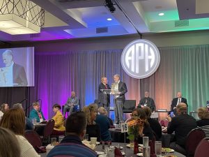 A man smiles as he accepts an award plaque from another man on a stage that is colorfully backlit and has the APH logo projected in the background. In front of the stage the two men are standing on is a crowd of people sitting at round tables.