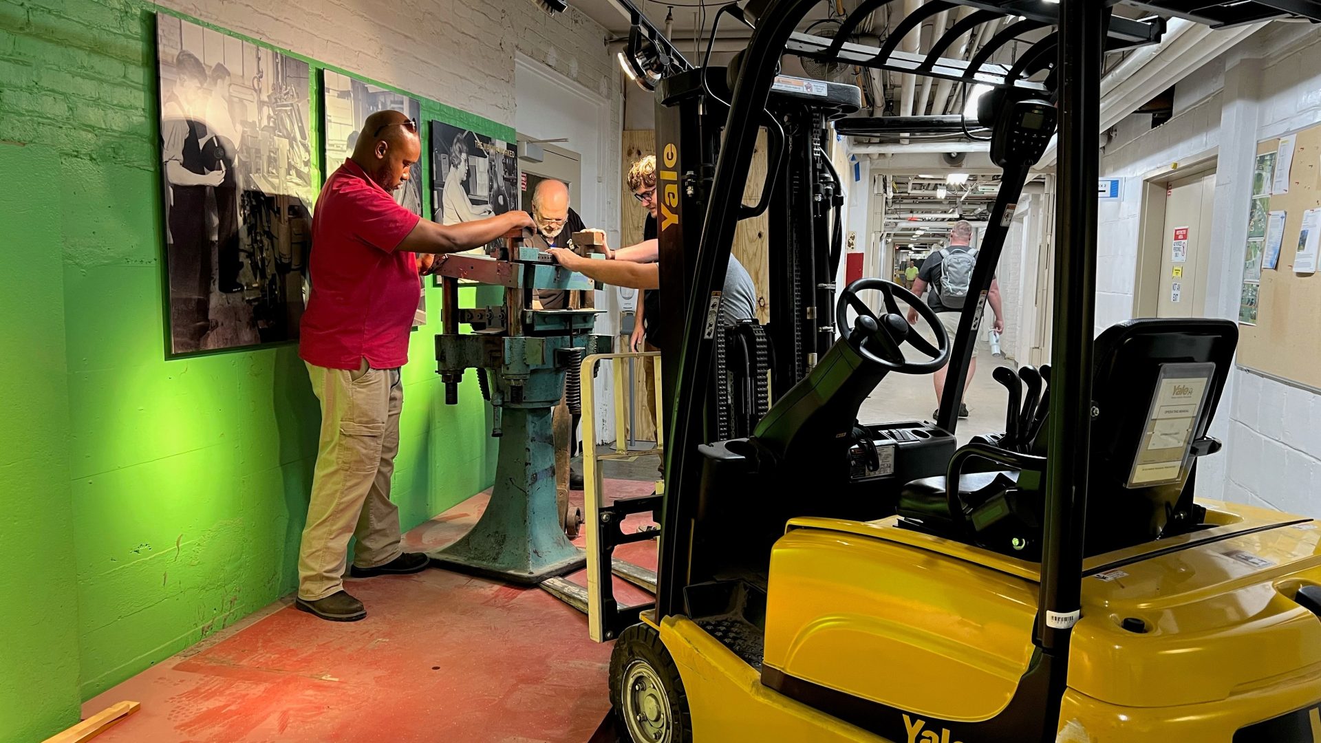 A yellow forklift fills the right foreground, its forks inserted beneath the base of a light blue steel record pressing machine, while three men steady the machine with their hands.