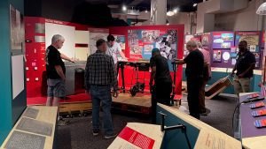 Seven men stand around a metal machine on a raised base in a partially dissembled museum exhibit. The men stare at the machine and gesture, as if figuring out their next move.