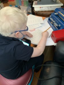 A young boy with light-colored hair writes on a piece of paper with simple arithmetic problems on it. On the surface he is working on, there is also an APH light-touch Perkins Brailler and a LEGO Braille Bricks box.
