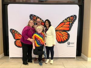 A smiling boy with light-colored hair wearing a tie-dye shirt stands with two women and a man in front of a photo backdrop with two sets of Monarch Butterfly wings on it. The APH logo and the Monarch logo are visible on the background in the bottom right hand corner.