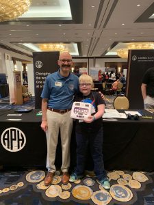 A smiling APH employee with a mustache and glasses poses with a young boy who has light-colored hair and glasses and is holding a Polly. They are in front of the APH booth in a convention hall.