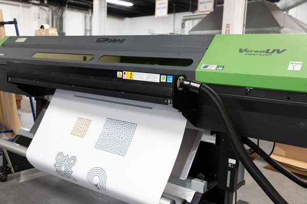 A Roland printing machine with a black and lime green body in an industrial room. The words “Roland” and “VersaUV Print & Cut” can be seen on the body, and a large white piece of paper with small mazes on it is coming out of the bottom of the machine.