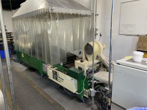 A long short green machine in an industrial room. Above the machine, thick clear plastic strips hang down from a metal hood to form a curtain over and around the machine.