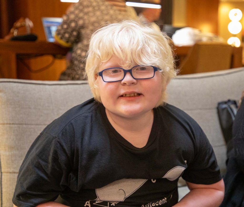A young boy with glasses and light-colored hair smiles as he poses for a photo.
