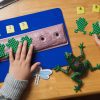 Two small hands attach the green tactile frog shapes to the log sitting on the blue storyboard with velcro stickers. Above each frog is a combination braille and large print number card that corresponds to the frog's place in line.