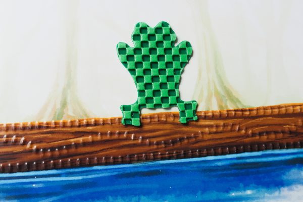 A green frog-shaped tactile piece with a checkerboard textured surface laid on an illustration of a log floating on blue water.