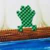 A green frog-shaped tactile piece with a checkerboard textured surface laid on an illustration of a log floating on blue water.