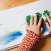 Two hands of a tactile reader on the open storybook. On the right page, the reader's hands sit on four green tactile frogs laid out on an illustration of a log floating on blue water. On the left page is a small illustration of a blue pond beneath braille and large print text.