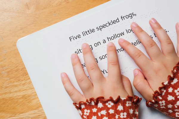 A close-up of two hands reading the braille on the first page of Five Little Speckled Frogs. Braille is embossed between lines of large print.