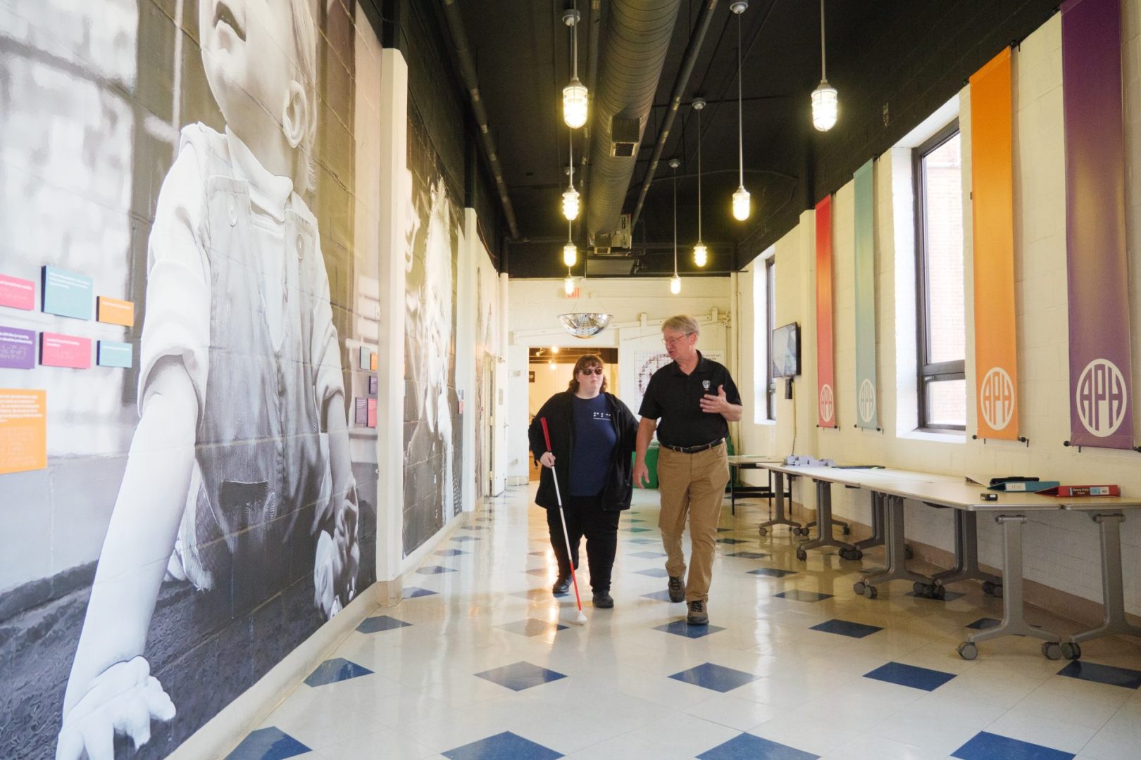 A man wearing a black shirt with the APH logo on it speaks to a young woman, who is wearing dark sunglasses and holding a white tipped cane, as he leads her down a hallway with a large black and white mural of a child on their right and a plain wall lined with banners displaying the APH logo in APH branding colors between tall windows on their left. There are also tables pushed against the wall on the left, and the light coming in from the windows reflects off the blue and white tiled floor.