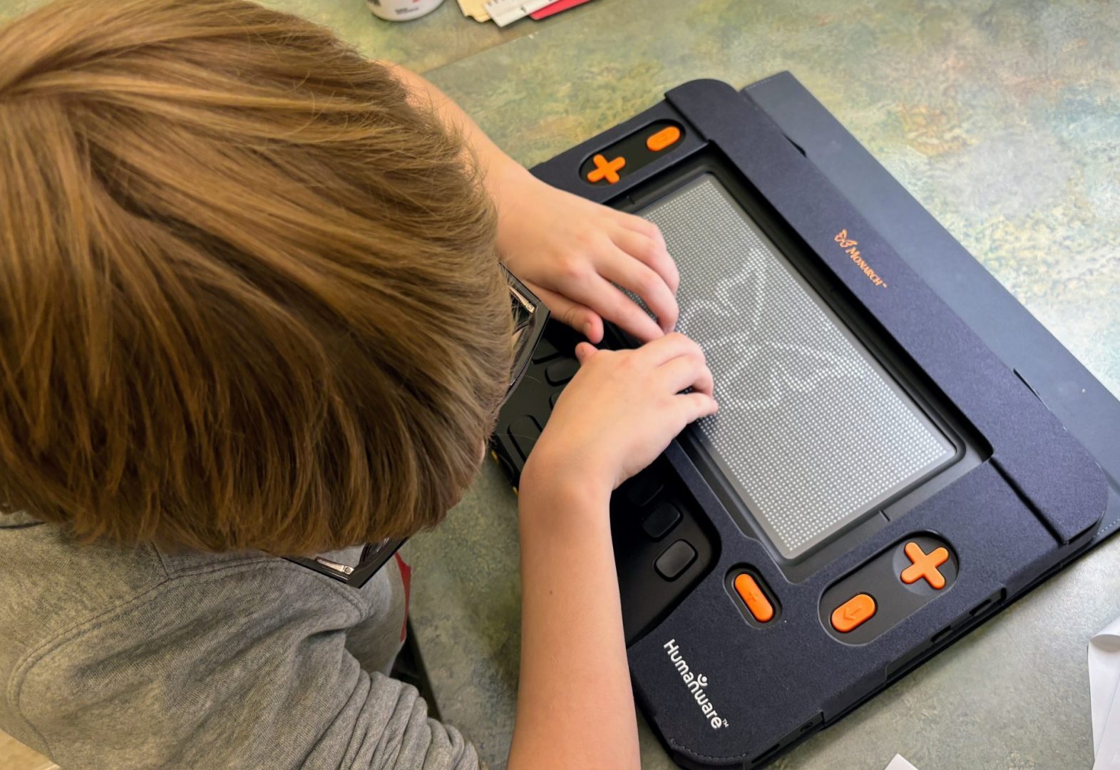 A young boy uses both of his hands to feel a tactile graphic of the butterfly featured in the Monarch logo on the Monarch’s 10 line by 32 cell refreshable braille display.