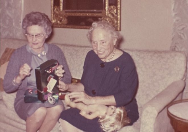 Two women sit on a sofa. At right, an older woman, Helen Keller, wearing a blue dress, has a child’s doll on her lap and seems to be accepting a wrapped present from the woman beside her. Helen has a big smile on her face and seems to be enjoying herself.